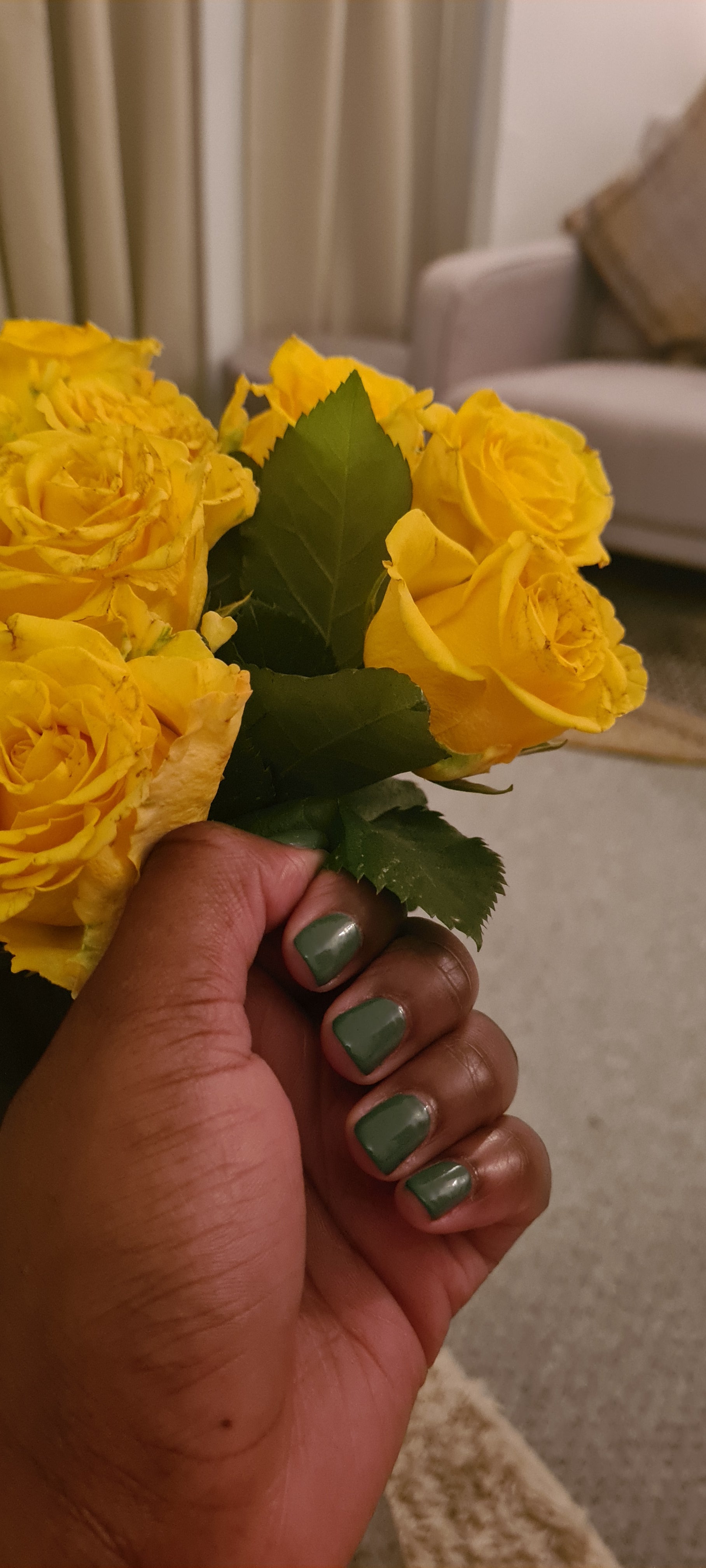 Swatch of olive green nail polish on nails, around yellow roses.